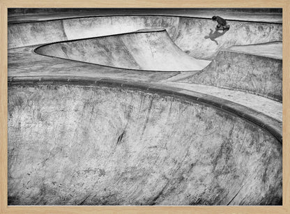 A high-angle, black and white photograph of a sprawling concrete skatepark with smooth, curving ramps. A single skateboarder is visible in the upper right, casting a distinct shadow on the ramp as they ride. The image is enclosed in a silver frame. Decor