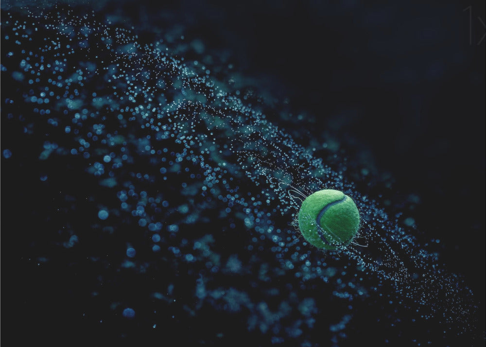 A high-speed photograph captures a spinning green tennis ball against a black background, with a dramatic spray of blue-lit water droplets flying off it in a cosmic-like spiral. Artwork