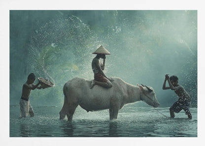 A serene photograph of three young boys playing in a river with a large water buffalo, one boy sits atop the animal while the others splash water, all set against a misty green backdrop and framed in silver. Print