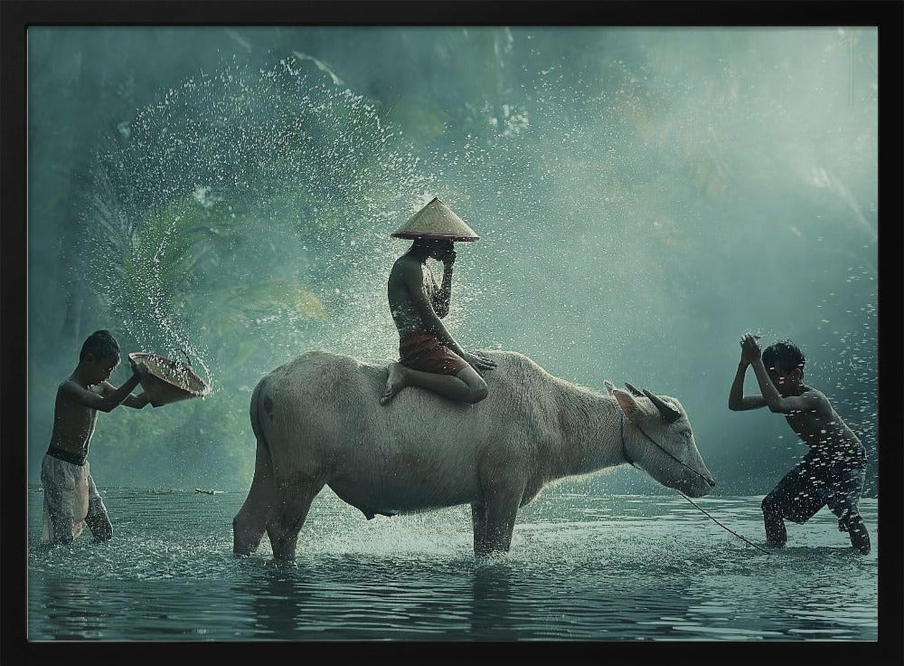 A serene photograph of three young boys playing in a river with a large water buffalo, one boy sits atop the animal while the others splash water, all set against a misty green backdrop and framed in silver. Print