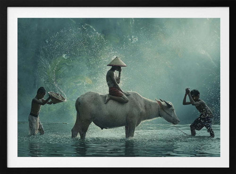 A serene photograph of three young boys playing in a river with a large water buffalo, one boy sits atop the animal while the others splash water, all set against a misty green backdrop and framed in silver. Print
