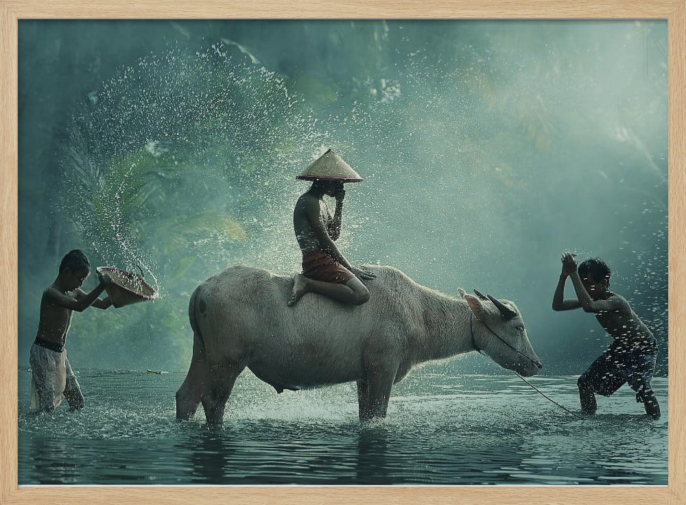 A serene photograph of three young boys playing in a river with a large water buffalo, one boy sits atop the animal while the others splash water, all set against a misty green backdrop and framed in silver. Print