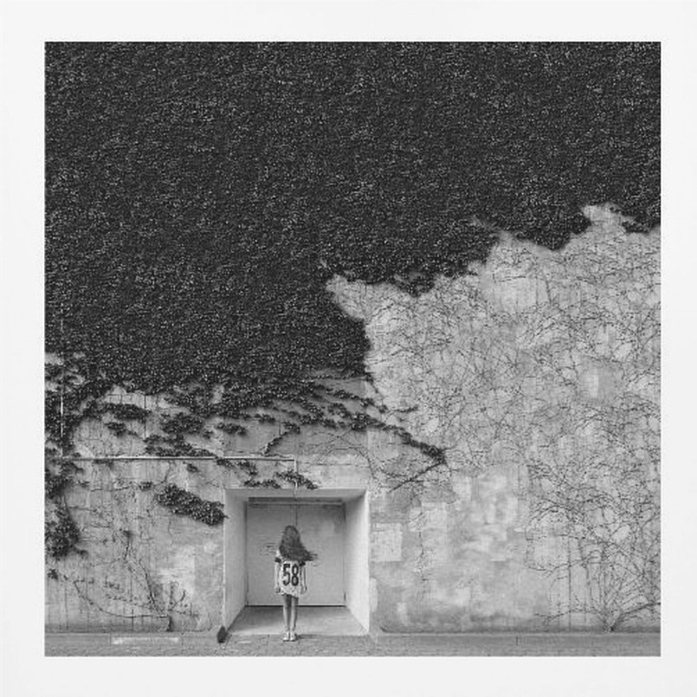 A framed, square black and white photograph showing a stark contrast on an ivy-covered wall. The top half is dense with dark leaves, while the bottom half is a lighter, textured wall. A young girl with long hair, seen from behind, stands in a recessed white doorway in the center. Wall Art