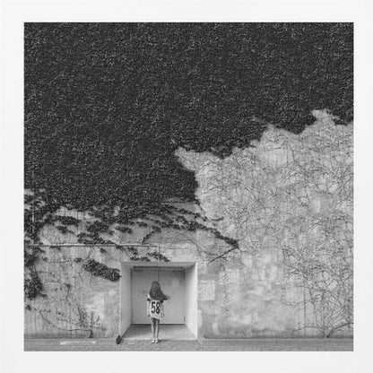 A framed, square black and white photograph showing a stark contrast on an ivy-covered wall. The top half is dense with dark leaves, while the bottom half is a lighter, textured wall. A young girl with long hair, seen from behind, stands in a recessed white doorway in the center. Wall Art