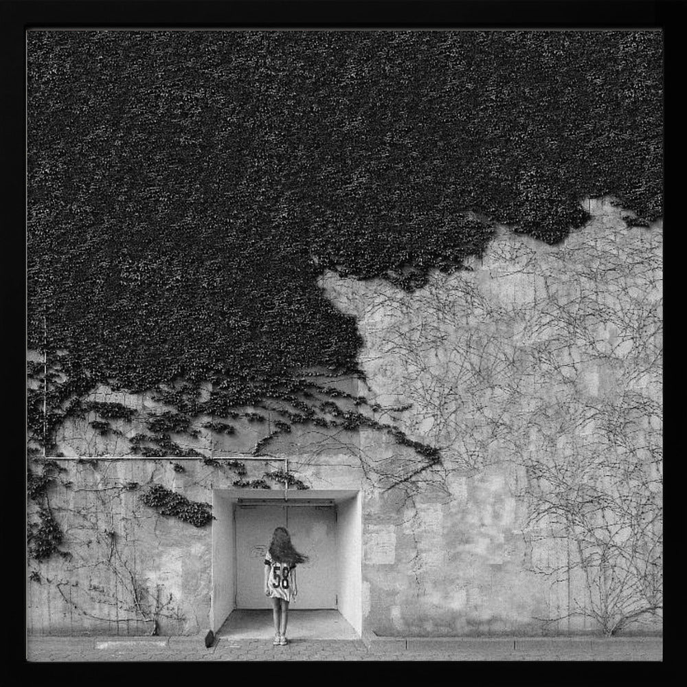 A framed, square black and white photograph showing a stark contrast on an ivy-covered wall. The top half is dense with dark leaves, while the bottom half is a lighter, textured wall. A young girl with long hair, seen from behind, stands in a recessed white doorway in the center. Wall Art