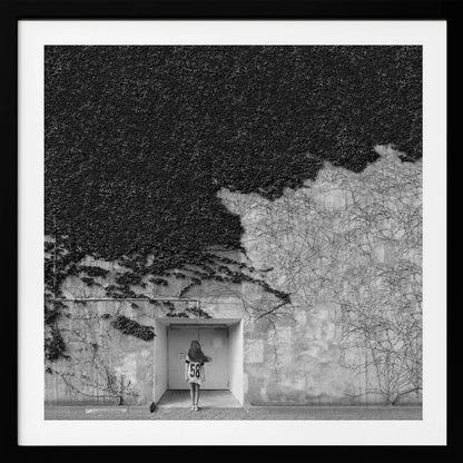 A framed, square black and white photograph showing a stark contrast on an ivy-covered wall. The top half is dense with dark leaves, while the bottom half is a lighter, textured wall. A young girl with long hair, seen from behind, stands in a recessed white doorway in the center. Wall Art