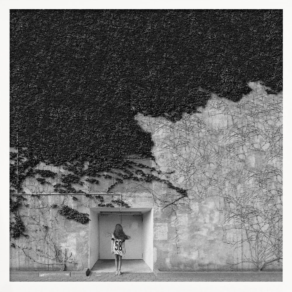 A framed, square black and white photograph showing a stark contrast on an ivy-covered wall. The top half is dense with dark leaves, while the bottom half is a lighter, textured wall. A young girl with long hair, seen from behind, stands in a recessed white doorway in the center. Wall Art