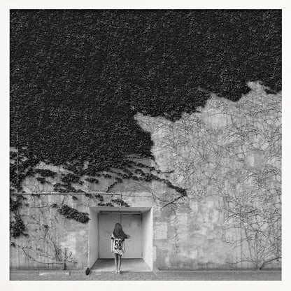 A framed, square black and white photograph showing a stark contrast on an ivy-covered wall. The top half is dense with dark leaves, while the bottom half is a lighter, textured wall. A young girl with long hair, seen from behind, stands in a recessed white doorway in the center. Wall Art