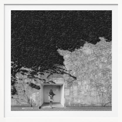 A framed, square black and white photograph showing a stark contrast on an ivy-covered wall. The top half is dense with dark leaves, while the bottom half is a lighter, textured wall. A young girl with long hair, seen from behind, stands in a recessed white doorway in the center. Wall Art