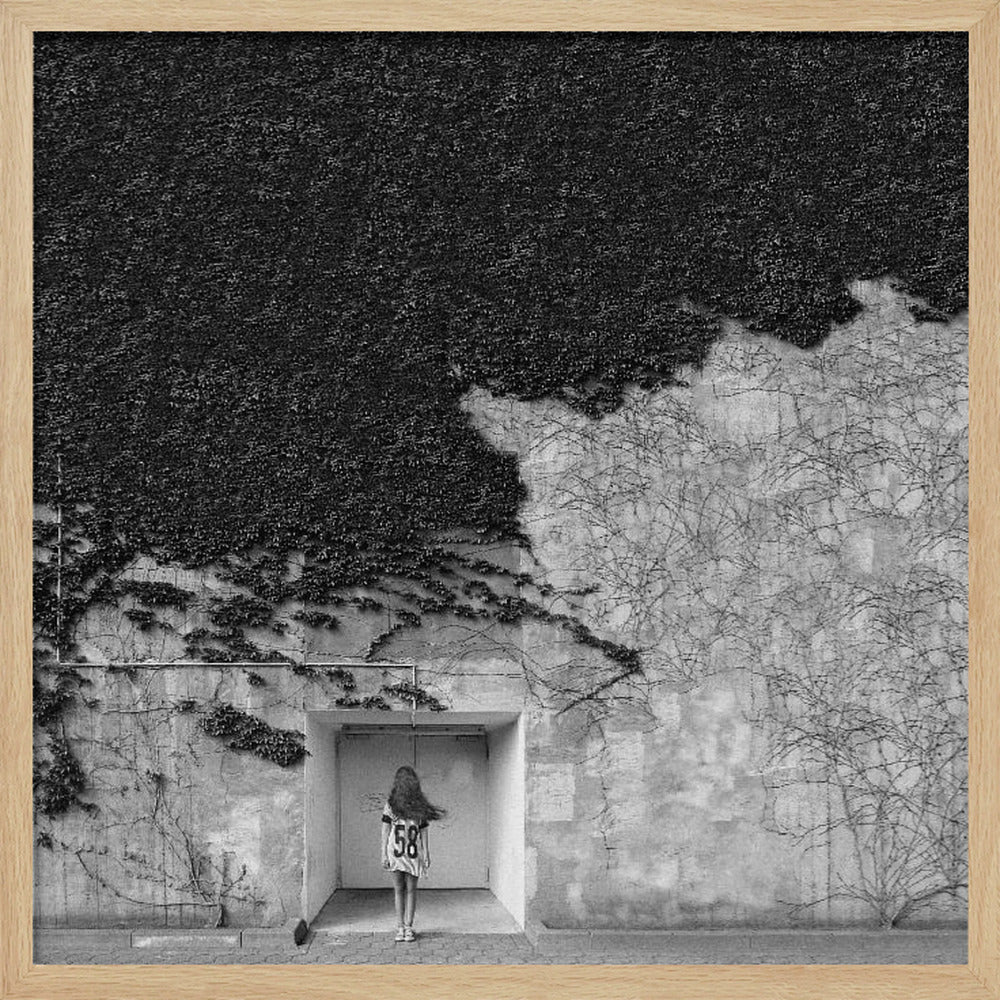 A framed, square black and white photograph showing a stark contrast on an ivy-covered wall. The top half is dense with dark leaves, while the bottom half is a lighter, textured wall. A young girl with long hair, seen from behind, stands in a recessed white doorway in the center. Wall Art