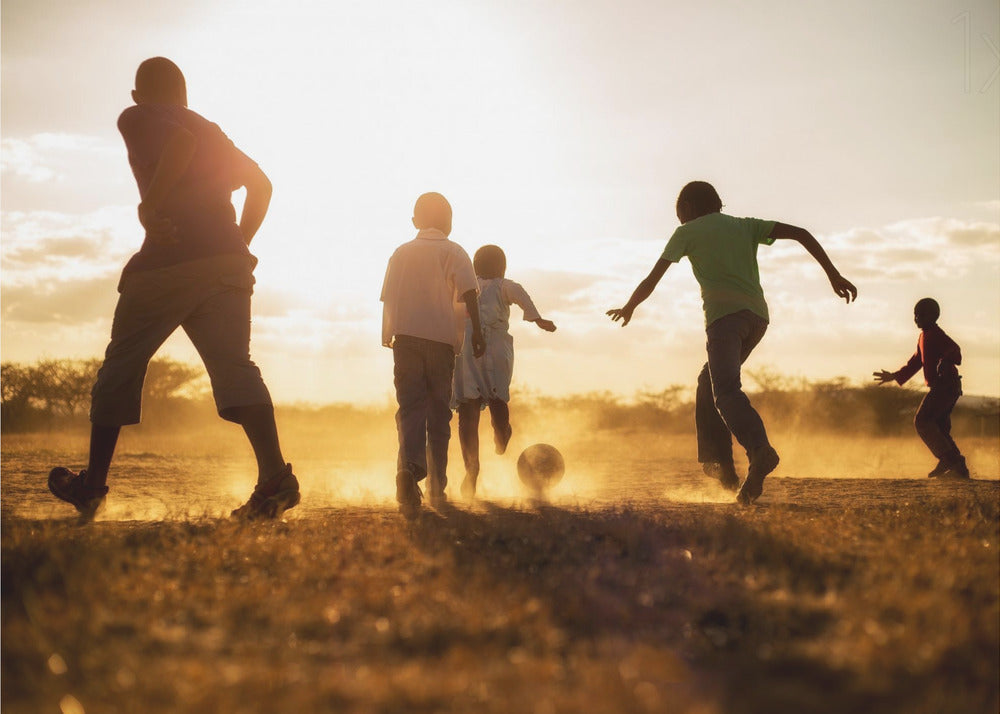 A low-angle, warmly lit photograph of five children silhouetted against a bright sunset as they play soccer on a dusty field, kicking up clouds of dust in the golden light. Decor