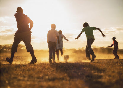A low-angle, warmly lit photograph of five children silhouetted against a bright sunset as they play soccer on a dusty field, kicking up clouds of dust in the golden light. Decor