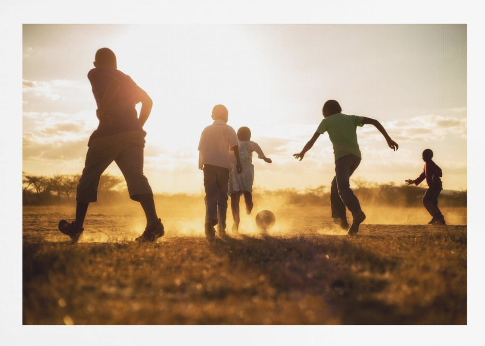 A low-angle, warmly lit photograph of five children silhouetted against a bright sunset as they play soccer on a dusty field, kicking up clouds of dust in the golden light. Decor