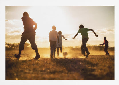 A low-angle, warmly lit photograph of five children silhouetted against a bright sunset as they play soccer on a dusty field, kicking up clouds of dust in the golden light. Decor