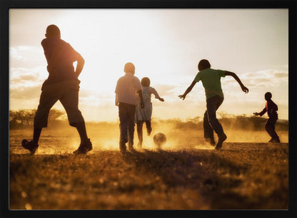 A low-angle, warmly lit photograph of five children silhouetted against a bright sunset as they play soccer on a dusty field, kicking up clouds of dust in the golden light. Decor