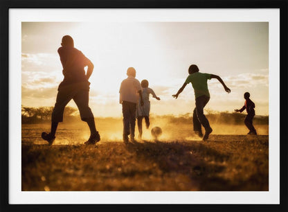 A low-angle, warmly lit photograph of five children silhouetted against a bright sunset as they play soccer on a dusty field, kicking up clouds of dust in the golden light. Decor