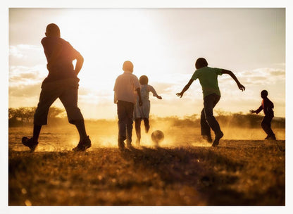 A low-angle, warmly lit photograph of five children silhouetted against a bright sunset as they play soccer on a dusty field, kicking up clouds of dust in the golden light. Decor