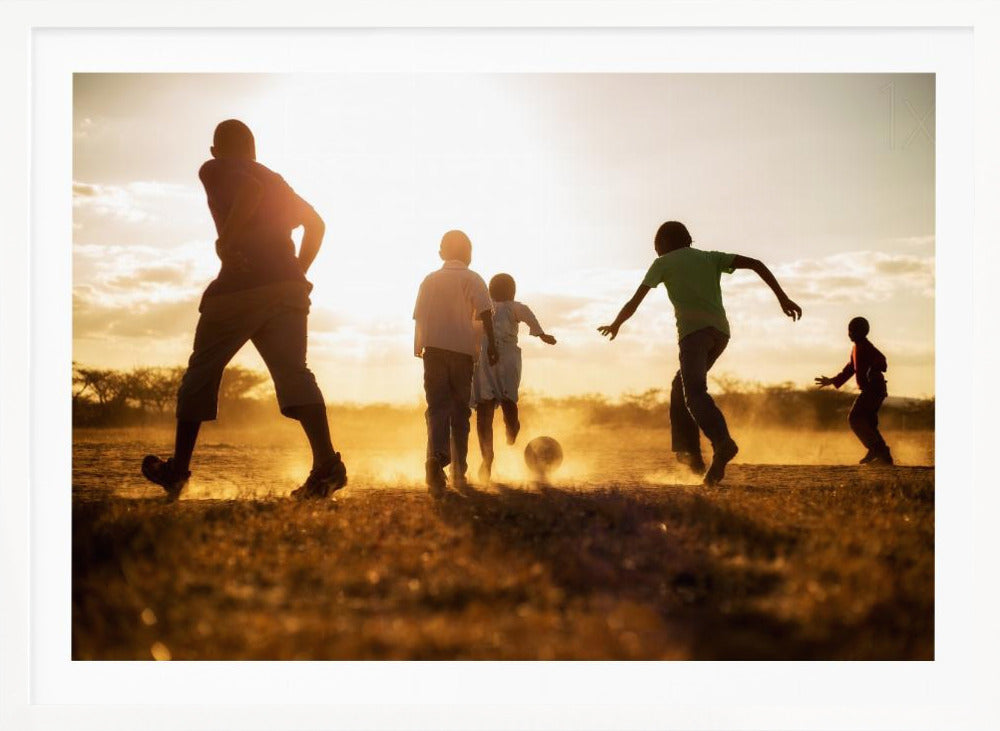A low-angle, warmly lit photograph of five children silhouetted against a bright sunset as they play soccer on a dusty field, kicking up clouds of dust in the golden light. Decor
