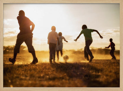 A low-angle, warmly lit photograph of five children silhouetted against a bright sunset as they play soccer on a dusty field, kicking up clouds of dust in the golden light. Decor