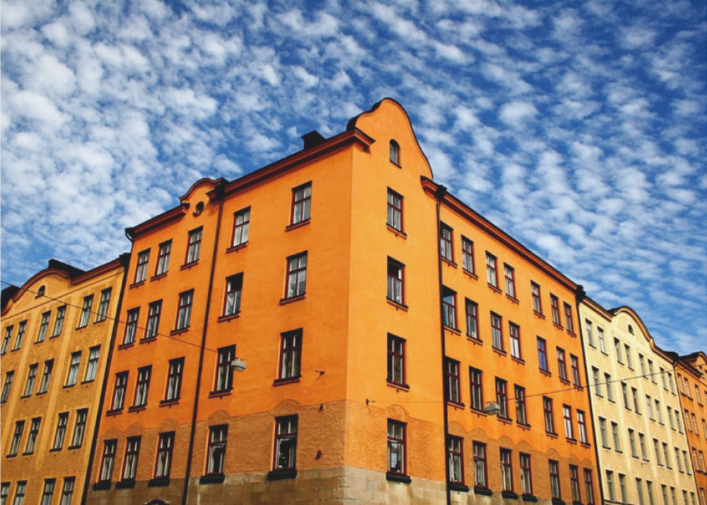 A low-angle shot of a vibrant orange apartment building against a dramatic blue sky filled with small, puffy white clouds. The building is on a corner, with another yellow-hued building visible next to it. The entire image is enclosed in a silver, brushed-metal style frame. Artwork