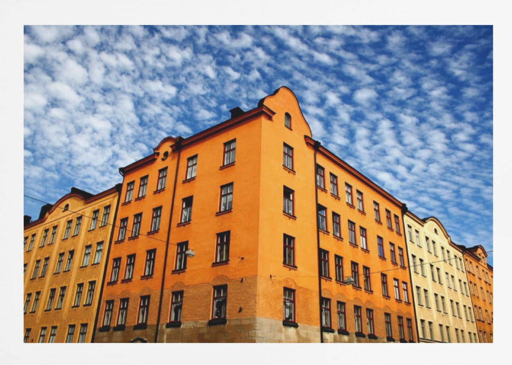 A low-angle shot of a vibrant orange apartment building against a dramatic blue sky filled with small, puffy white clouds. The building is on a corner, with another yellow-hued building visible next to it. The entire image is enclosed in a silver, brushed-metal style frame. Artwork