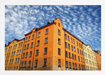 A low-angle shot of a vibrant orange apartment building against a dramatic blue sky filled with small, puffy white clouds. The building is on a corner, with another yellow-hued building visible next to it. The entire image is enclosed in a silver, brushed-metal style frame. Artwork