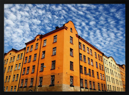 A low-angle shot of a vibrant orange apartment building against a dramatic blue sky filled with small, puffy white clouds. The building is on a corner, with another yellow-hued building visible next to it. The entire image is enclosed in a silver, brushed-metal style frame. Artwork