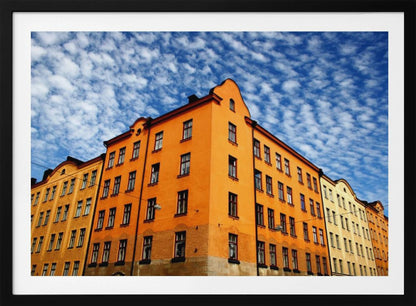 A low-angle shot of a vibrant orange apartment building against a dramatic blue sky filled with small, puffy white clouds. The building is on a corner, with another yellow-hued building visible next to it. The entire image is enclosed in a silver, brushed-metal style frame. Artwork