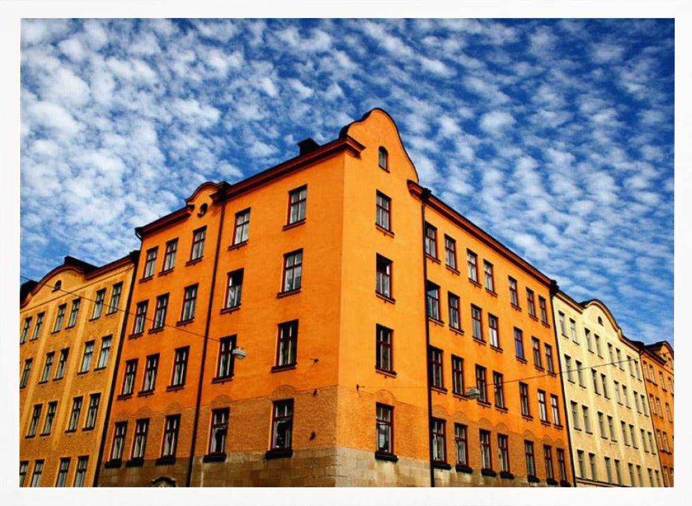 A low-angle shot of a vibrant orange apartment building against a dramatic blue sky filled with small, puffy white clouds. The building is on a corner, with another yellow-hued building visible next to it. The entire image is enclosed in a silver, brushed-metal style frame. Artwork