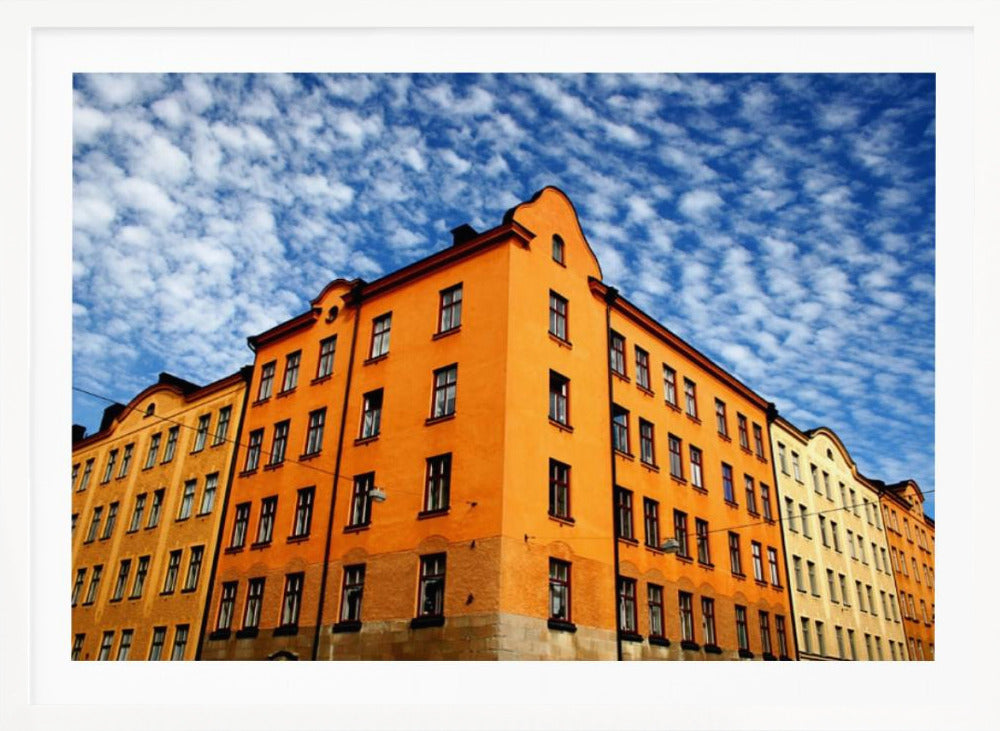 A low-angle shot of a vibrant orange apartment building against a dramatic blue sky filled with small, puffy white clouds. The building is on a corner, with another yellow-hued building visible next to it. The entire image is enclosed in a silver, brushed-metal style frame. Artwork