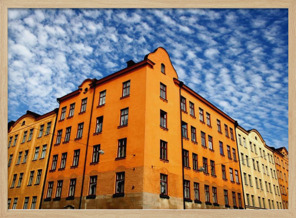 A low-angle shot of a vibrant orange apartment building against a dramatic blue sky filled with small, puffy white clouds. The building is on a corner, with another yellow-hued building visible next to it. The entire image is enclosed in a silver, brushed-metal style frame. Artwork