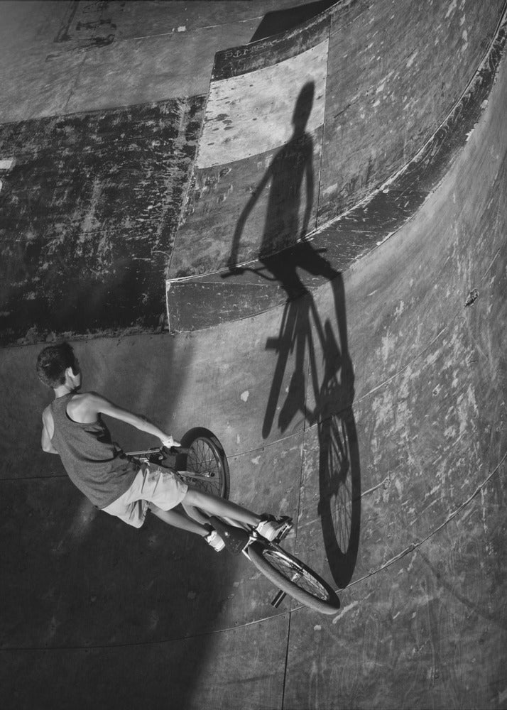 A dramatic black and white high-angle photograph of a BMX rider in a skate park. The rider is captured in motion on a curved ramp, while their long, stark shadow is cast on the worn, textured surface ahead, creating a powerful visual contrast. Poster
