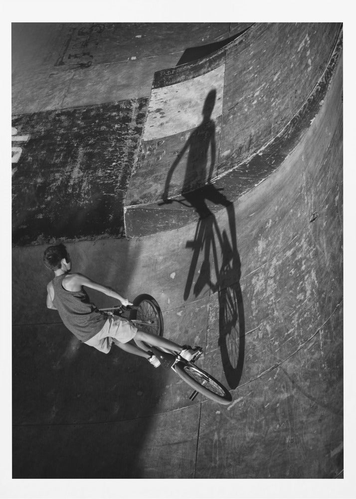 A dramatic black and white high-angle photograph of a BMX rider in a skate park. The rider is captured in motion on a curved ramp, while their long, stark shadow is cast on the worn, textured surface ahead, creating a powerful visual contrast. Poster