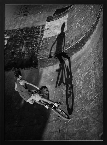 A dramatic black and white high-angle photograph of a BMX rider in a skate park. The rider is captured in motion on a curved ramp, while their long, stark shadow is cast on the worn, textured surface ahead, creating a powerful visual contrast. Poster
