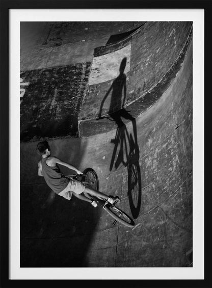 A dramatic black and white high-angle photograph of a BMX rider in a skate park. The rider is captured in motion on a curved ramp, while their long, stark shadow is cast on the worn, textured surface ahead, creating a powerful visual contrast. Poster