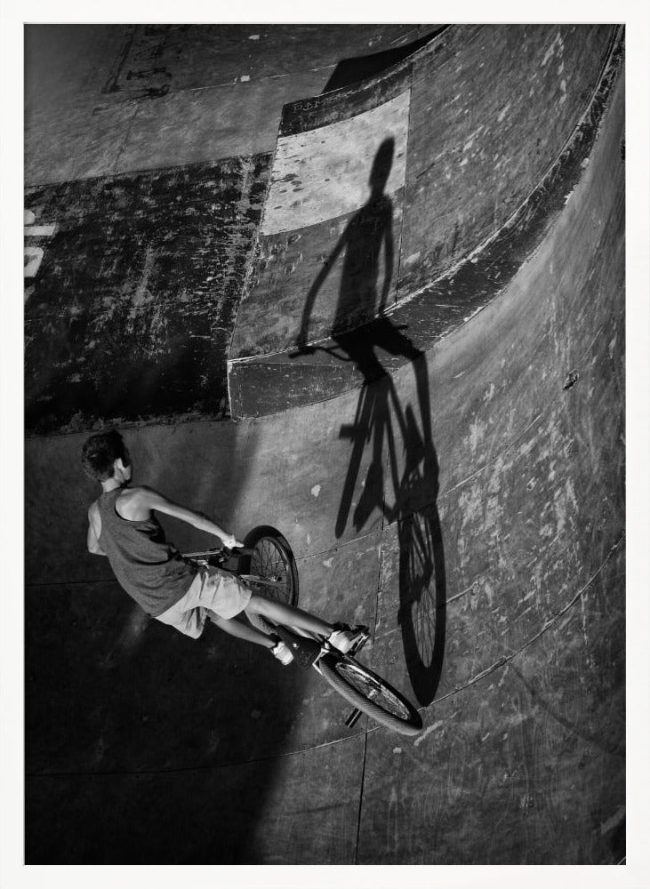 A dramatic black and white high-angle photograph of a BMX rider in a skate park. The rider is captured in motion on a curved ramp, while their long, stark shadow is cast on the worn, textured surface ahead, creating a powerful visual contrast. Poster