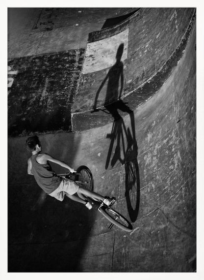 A dramatic black and white high-angle photograph of a BMX rider in a skate park. The rider is captured in motion on a curved ramp, while their long, stark shadow is cast on the worn, textured surface ahead, creating a powerful visual contrast. Poster