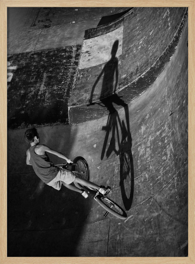 A dramatic black and white high-angle photograph of a BMX rider in a skate park. The rider is captured in motion on a curved ramp, while their long, stark shadow is cast on the worn, textured surface ahead, creating a powerful visual contrast. Poster
