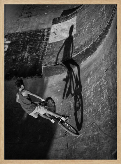 A dramatic black and white high-angle photograph of a BMX rider in a skate park. The rider is captured in motion on a curved ramp, while their long, stark shadow is cast on the worn, textured surface ahead, creating a powerful visual contrast. Poster