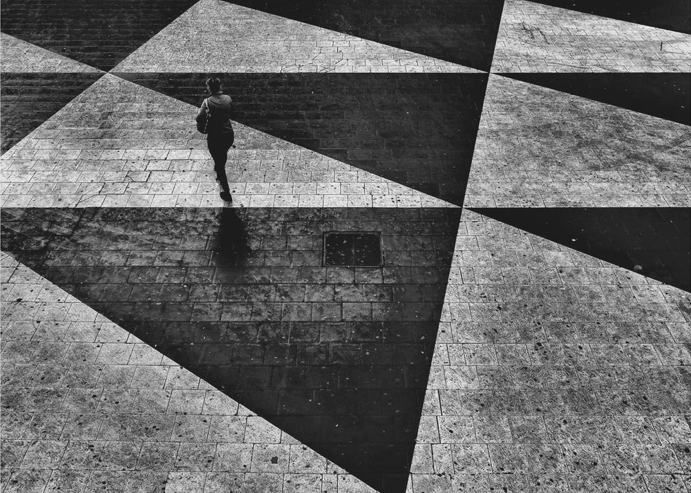 A dramatic, high-contrast black and white overhead photograph of a person walking across a paved ground with a striking geometric pattern of light and dark triangles created by strong shadows. The lone figure casts a long shadow, enhancing the minimalist and abstract composition, which is enclosed in a grey frame. Print