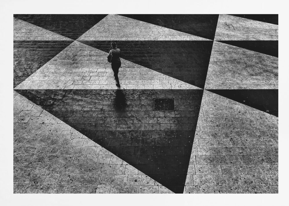 A dramatic, high-contrast black and white overhead photograph of a person walking across a paved ground with a striking geometric pattern of light and dark triangles created by strong shadows. The lone figure casts a long shadow, enhancing the minimalist and abstract composition, which is enclosed in a grey frame. Print