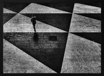 A dramatic, high-contrast black and white overhead photograph of a person walking across a paved ground with a striking geometric pattern of light and dark triangles created by strong shadows. The lone figure casts a long shadow, enhancing the minimalist and abstract composition, which is enclosed in a grey frame. Print