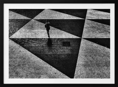 A dramatic, high-contrast black and white overhead photograph of a person walking across a paved ground with a striking geometric pattern of light and dark triangles created by strong shadows. The lone figure casts a long shadow, enhancing the minimalist and abstract composition, which is enclosed in a grey frame. Print
