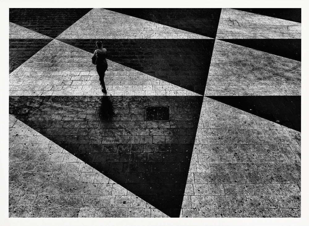 A dramatic, high-contrast black and white overhead photograph of a person walking across a paved ground with a striking geometric pattern of light and dark triangles created by strong shadows. The lone figure casts a long shadow, enhancing the minimalist and abstract composition, which is enclosed in a grey frame. Print