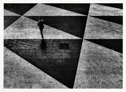 A dramatic, high-contrast black and white overhead photograph of a person walking across a paved ground with a striking geometric pattern of light and dark triangles created by strong shadows. The lone figure casts a long shadow, enhancing the minimalist and abstract composition, which is enclosed in a grey frame. Print