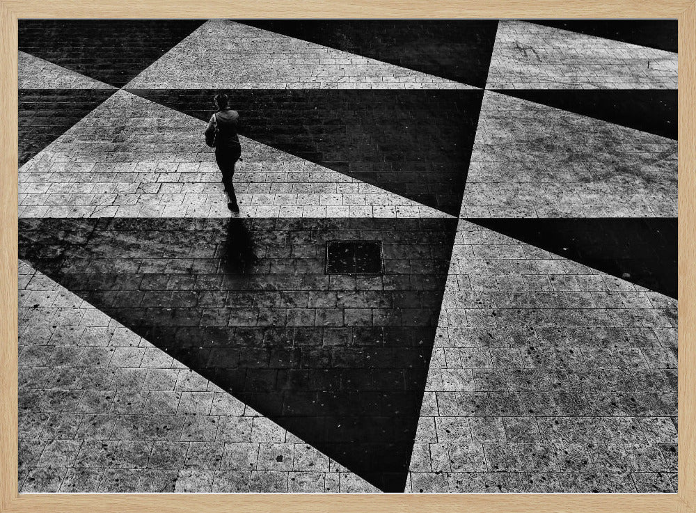 A dramatic, high-contrast black and white overhead photograph of a person walking across a paved ground with a striking geometric pattern of light and dark triangles created by strong shadows. The lone figure casts a long shadow, enhancing the minimalist and abstract composition, which is enclosed in a grey frame. Print