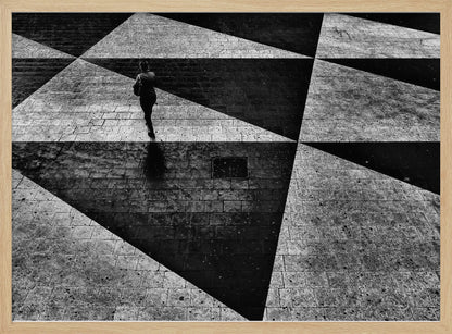 A dramatic, high-contrast black and white overhead photograph of a person walking across a paved ground with a striking geometric pattern of light and dark triangles created by strong shadows. The lone figure casts a long shadow, enhancing the minimalist and abstract composition, which is enclosed in a grey frame. Print