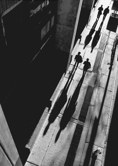 A dramatic high-angle, black and white photograph of several people walking on a sunlit sidewalk, casting long, stark shadows that stretch diagonally across the frame, contrasting with the deep black shadows of the adjacent building. Decor