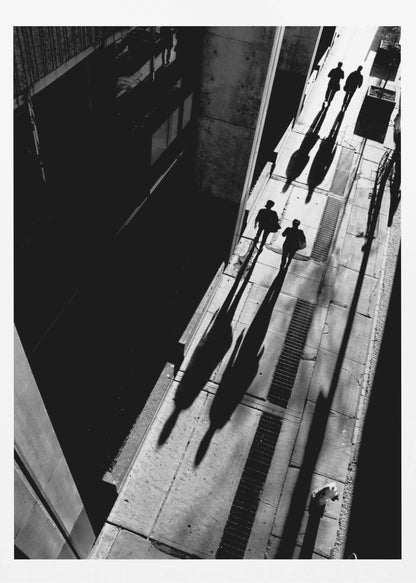 A dramatic high-angle, black and white photograph of several people walking on a sunlit sidewalk, casting long, stark shadows that stretch diagonally across the frame, contrasting with the deep black shadows of the adjacent building. Decor