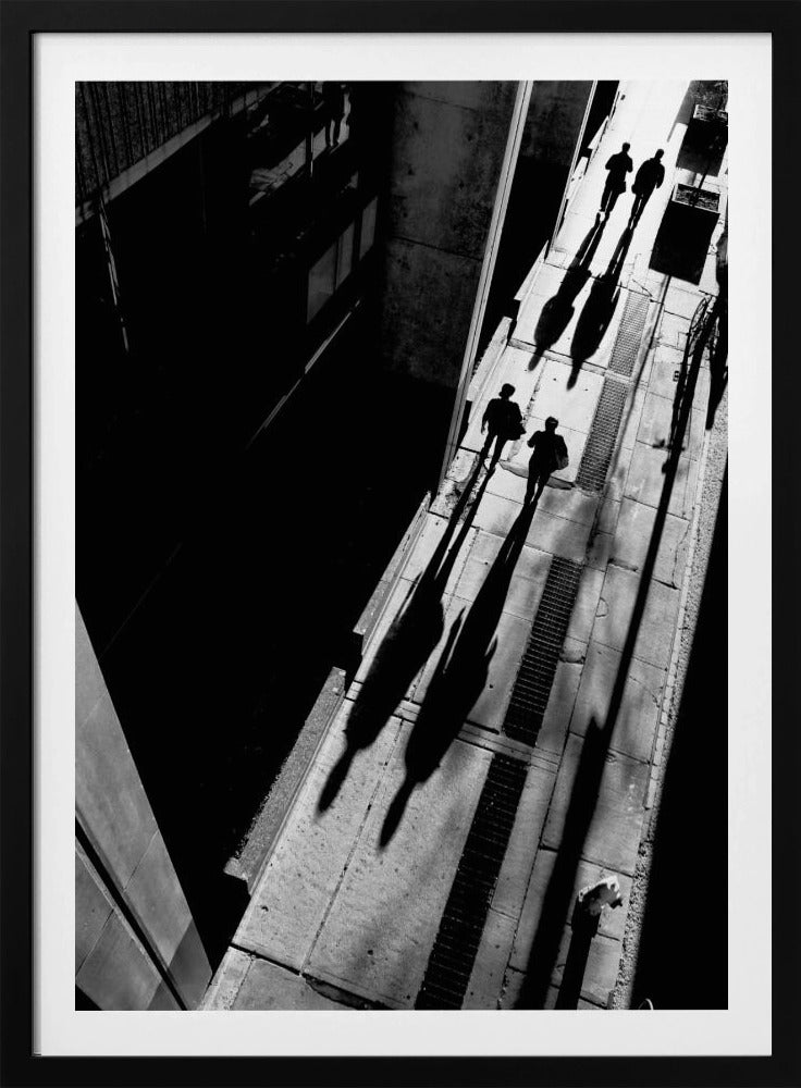 A dramatic high-angle, black and white photograph of several people walking on a sunlit sidewalk, casting long, stark shadows that stretch diagonally across the frame, contrasting with the deep black shadows of the adjacent building. Decor